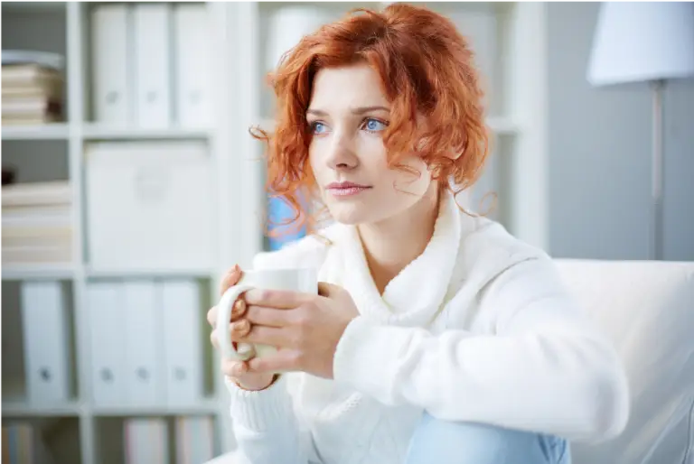 Anxious woman sitting on couch in white sweater holding a coffee mug trying to overcome social anxiety
